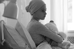 Child sitting in bed wearing a headwrap and holding a teddy bear.