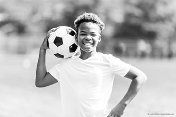 A young boy wearing a white shirt is smiling at the camera while holding a soccer ball.