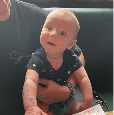 A smiling baby sits on an adult’s lap at a restaurant, reaching toward food on the table. The baby looks healthy and alert, dressed in a navy onesie with planets, contrasting with a hospital scene visible in the background through a window.