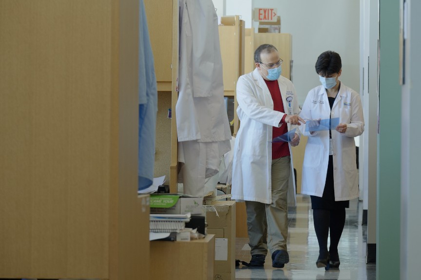 Dr. Fouladi is wearing a white coat and surgical mask. She's walking down a hallway and collaborating with one of her colleagues, also pictured in a white coat and surgical mask.
