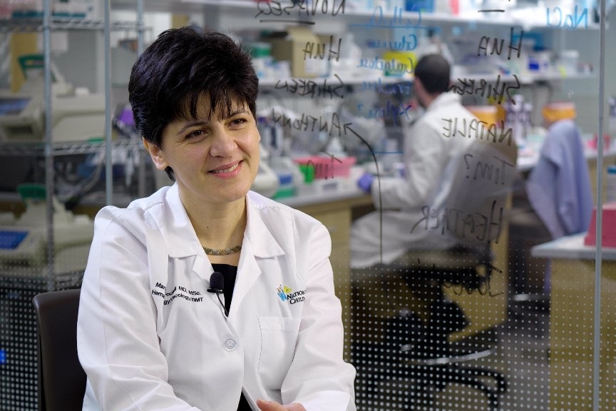 Dr. Fouladi is wearing a white coat and is seated in a laboratory setting.