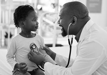 A male medical provider is listening to his patient's heart beat with a stethoscope. The patient is a little girl wearing a white shirt and looking to the side.
