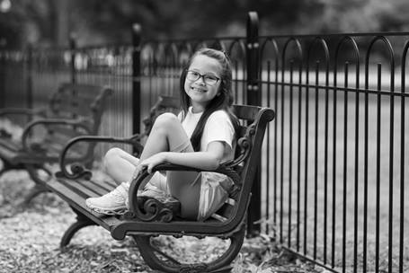 An elementary-aged girl is smiling, sitting on a park bench. She's wearing glasses and a backpack. A metal fence is in the background behind her.