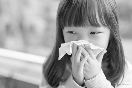An elementary aged girl with dark brown hair and bangs holds a tissue up to her nose.