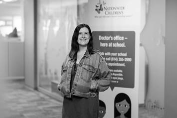 Bridget is wearing a jean jacket, with her hands resting in her pockets. She's smiling, standing in an office setting with a school health banner behind her.