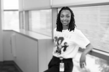 Dee is wearing a white graphic tee and dark pants. She's smiling, leaning against a wall of windows.