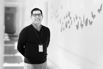 Dr. Beltran is standing in a hallway with butterfly graphics on the wall. He's smiling, wearing a dark sweater and glasses.