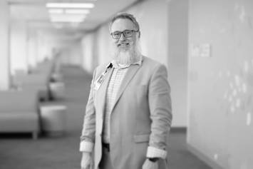 John is smiling, standing in a hallway with a row of chairs behind him. He's wearing glasses and a light colored suit jacket.