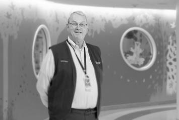 Tom is smiling with arms behind his back. He's wearing a volunteer vest with leaf graphics on the wall behind him.