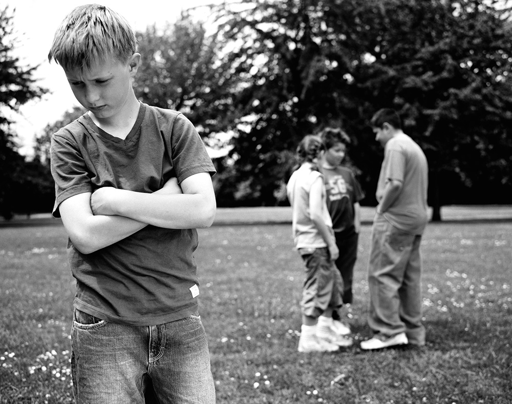 a young boy with his arms crossed looking sad. Group of 3 kids behind talking about him