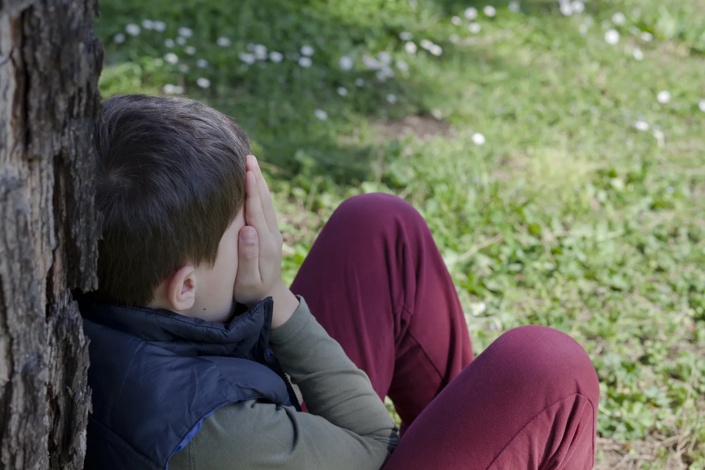 child sitting against a tree with their hands covering their face