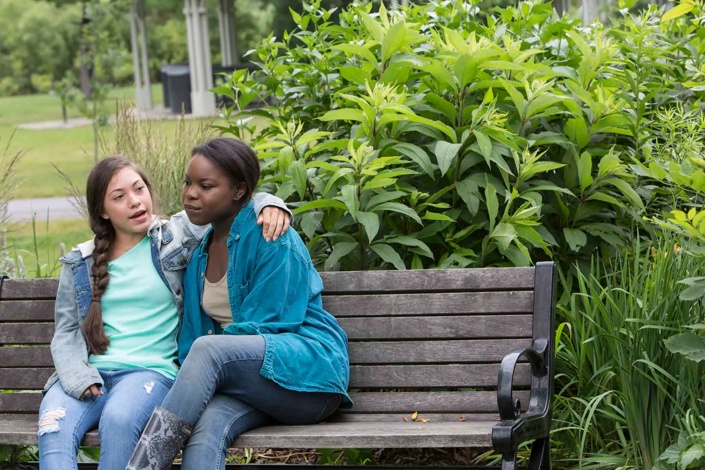 A teenage girl sitting on a park bench with her arm around her friend.