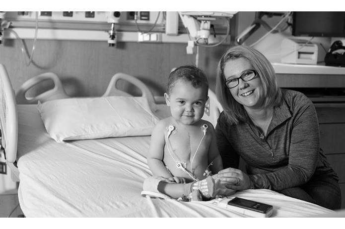 baby sitting on hospital bed with mom leaning on the bed smiling at the camera