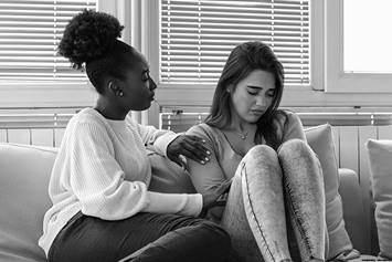 Two girls sitting on a couch. One girl consoling the other.