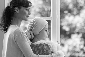Child with cancer wearing headscarf, looking out the window with their mom