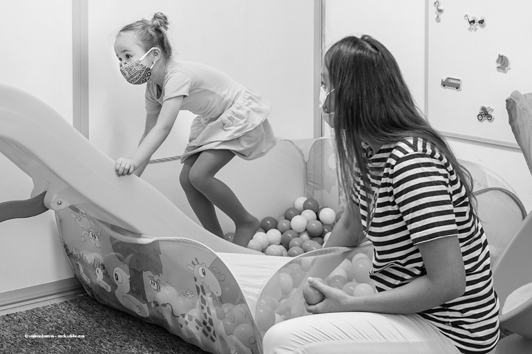 Little girl wearing a mask while playing in a ball bit while mom watches.