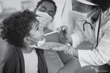doctor, wearing a face mask, examining patients mouth, while parent looks on.