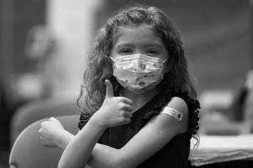 Little girl wearing a mask with a band-aid on her arm, giving a thumbs up.