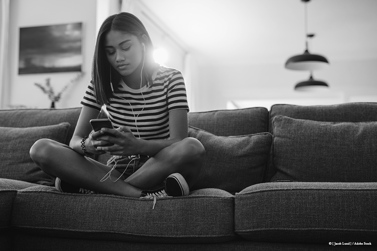 girl siting on a couch, looking at her phone and wearing ear buds.