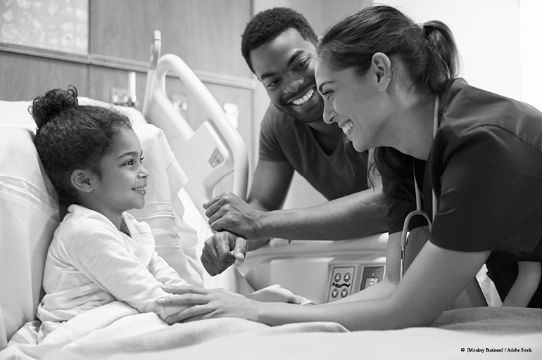 child sitting in a hospital bed with two adults looking over her.