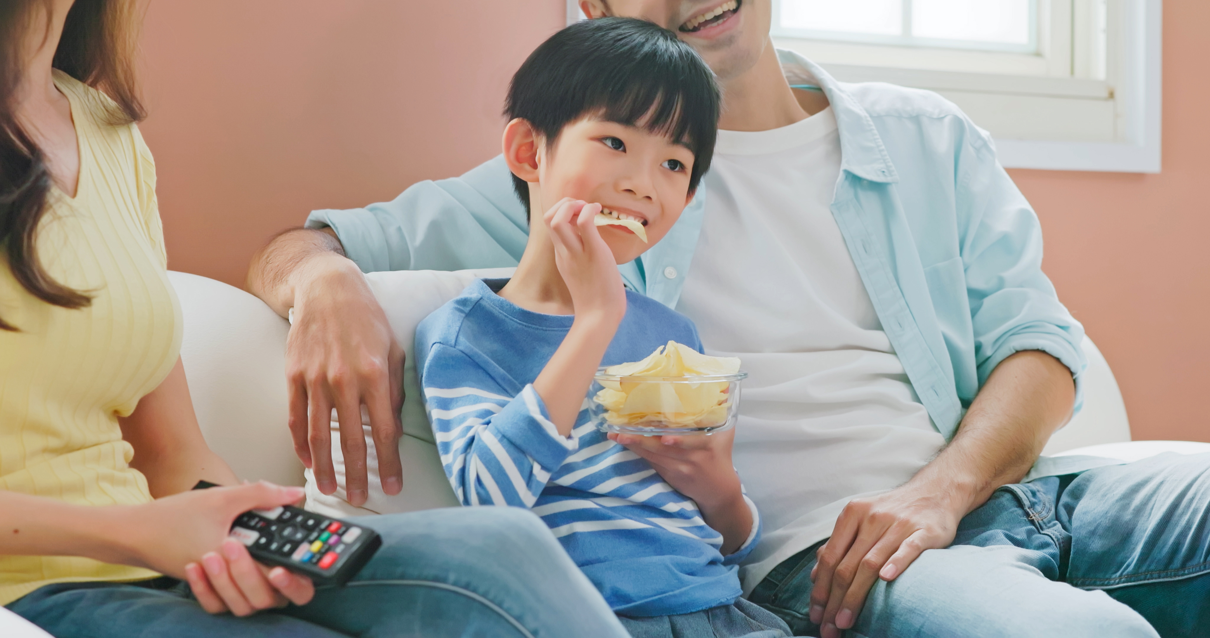 Young boy eating chips on the couch, sitting between his parents. 
