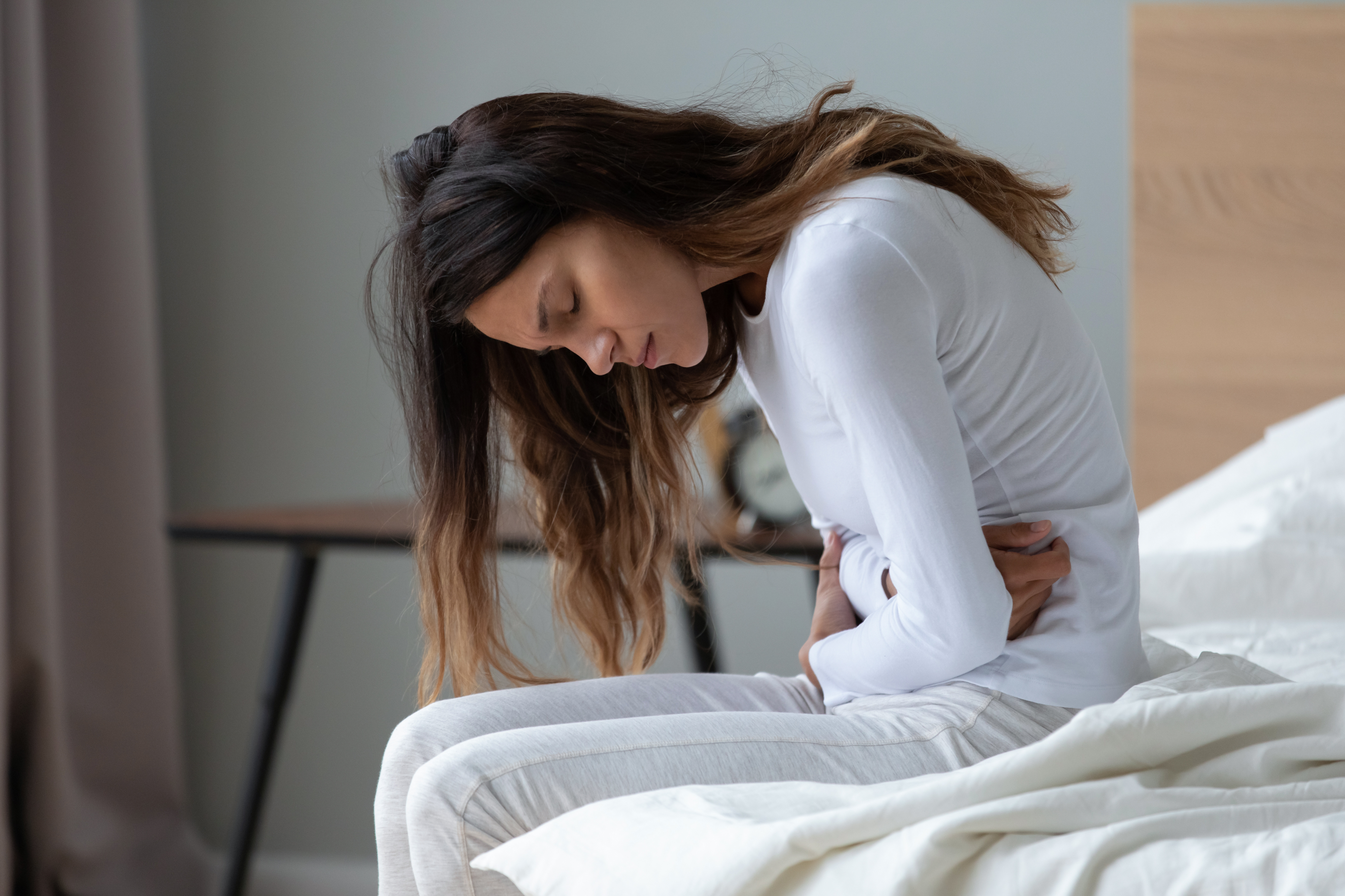young woman sitting on the edge of a bed, clutching her stomach in pain