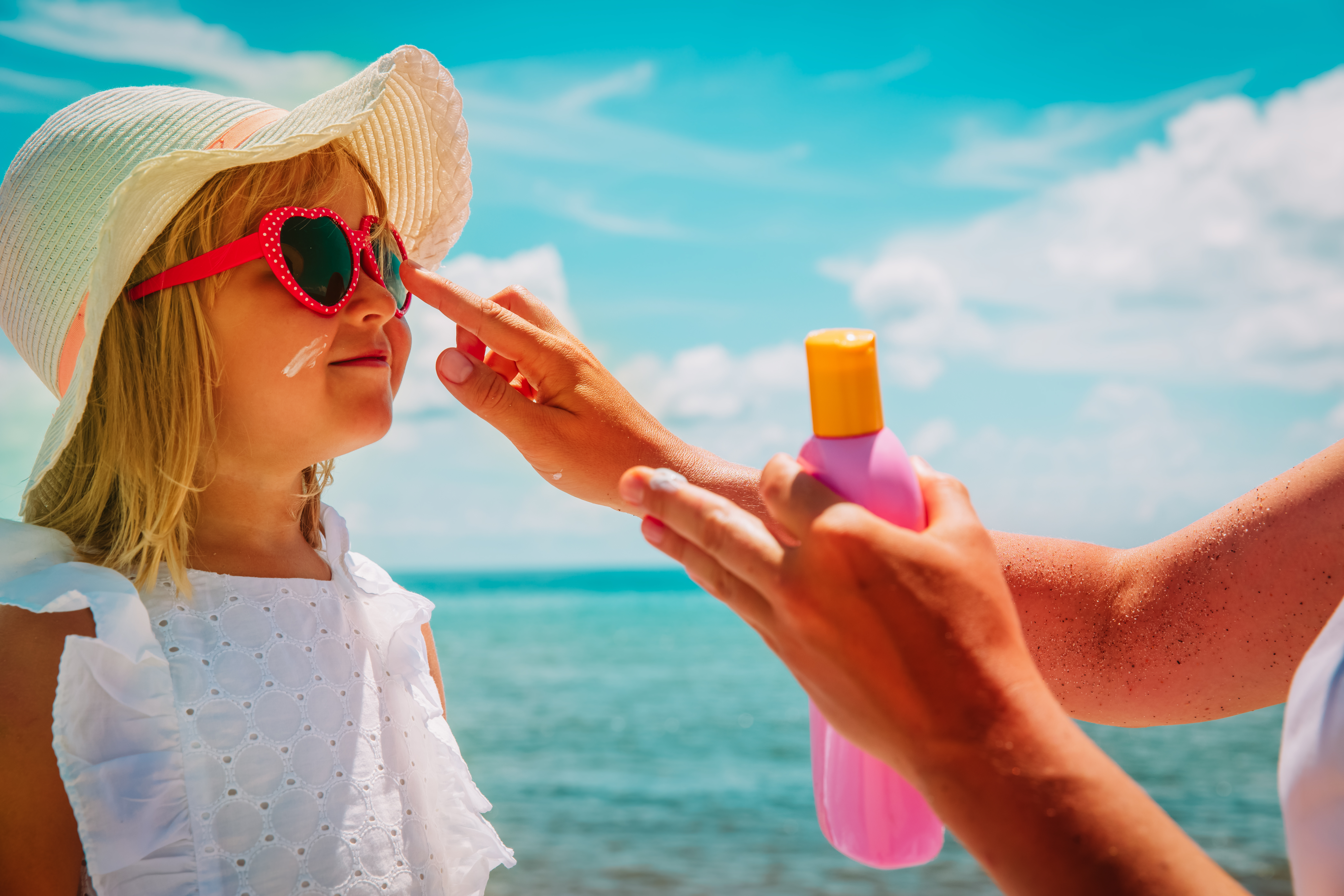 young girl with sunglasses and hat receiving help putting on sunscreen