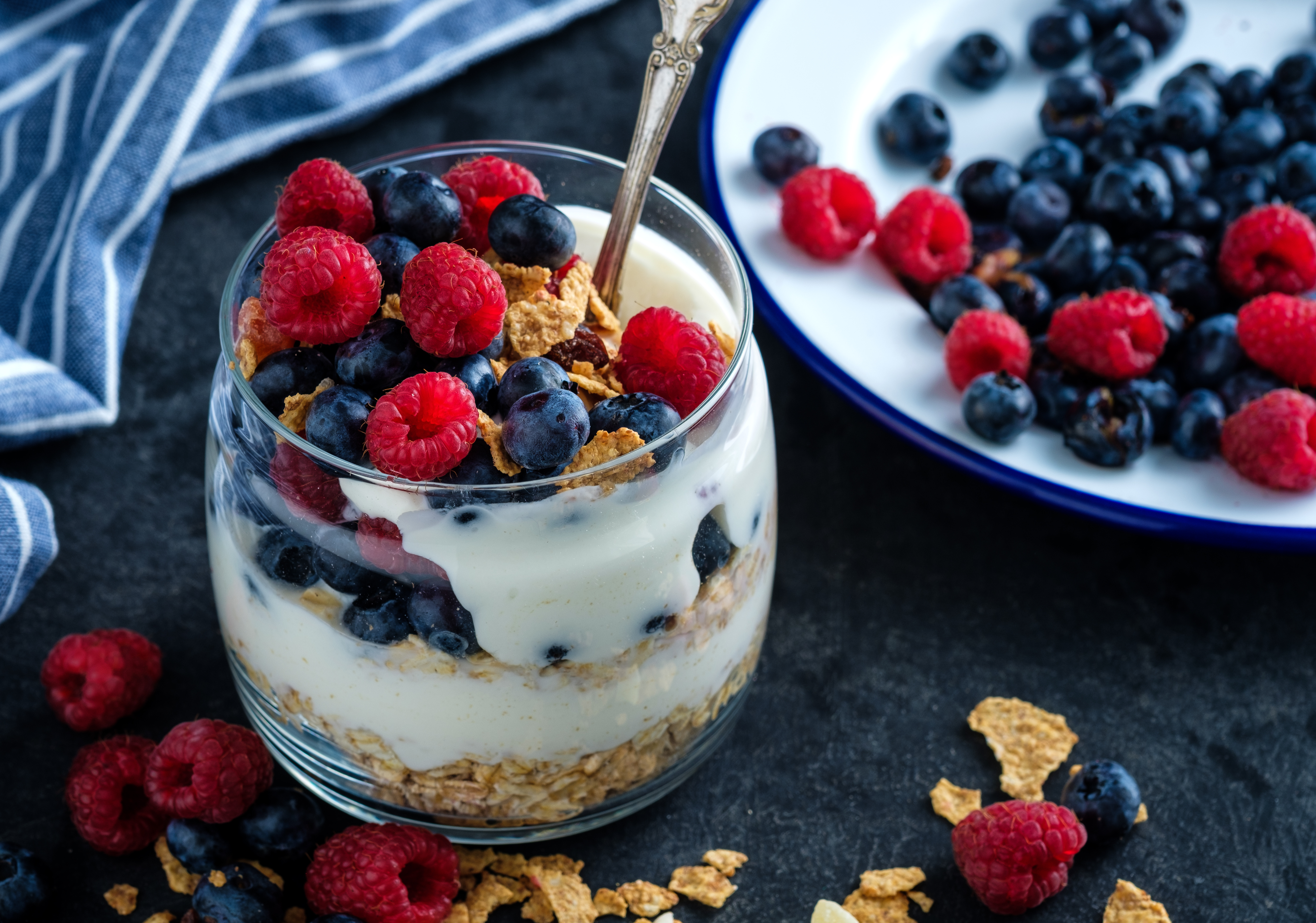 A glass filled with layers of granola, yogurt, blueberries, and raspberries sits on a dark surface with a spoon inside. Additional berries are scattered around, with a striped napkin and a plate of fruit in the background.