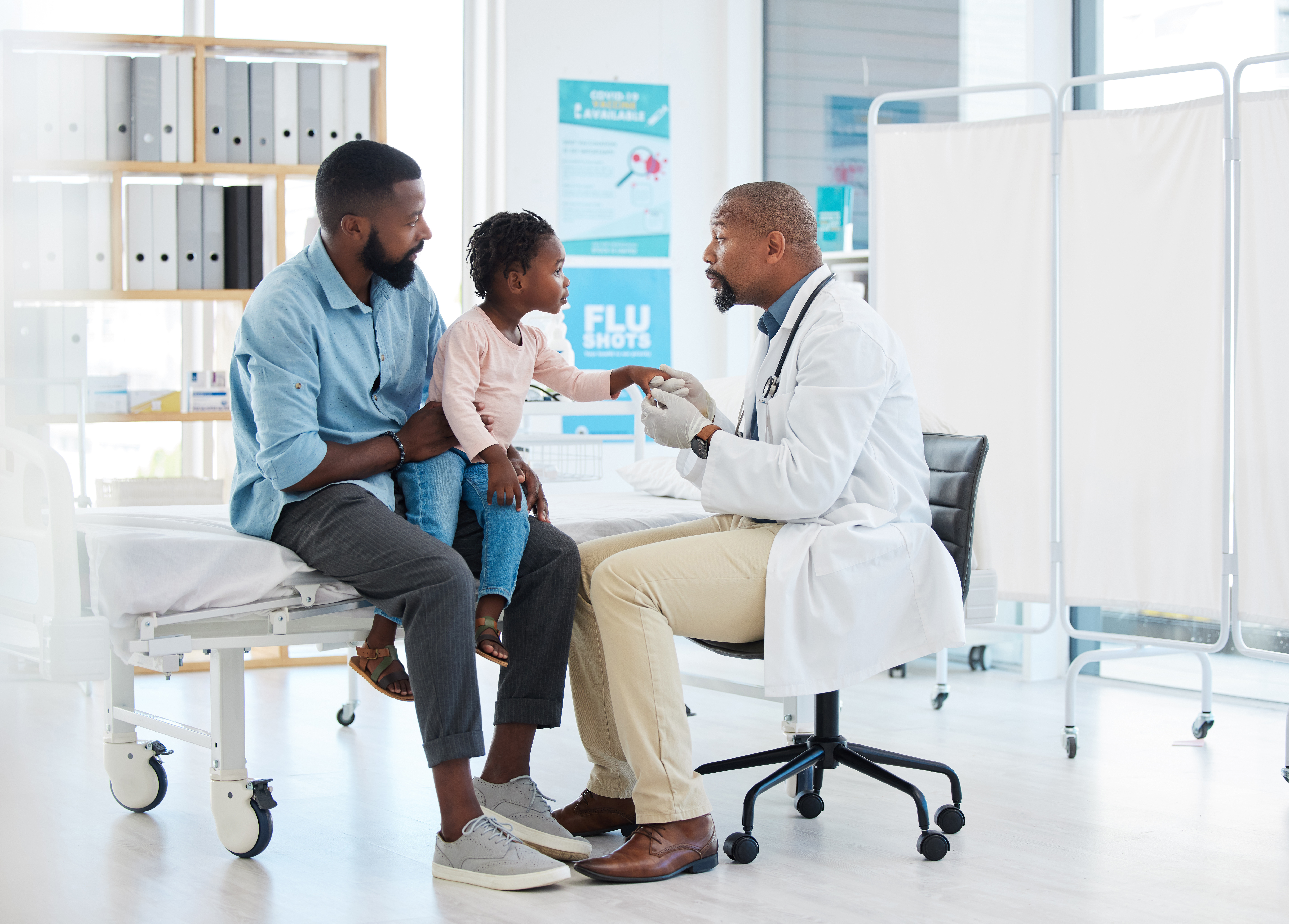 A provider is holding a young patient's hand, while his father holds him. There is a sign for flu shots in the background.