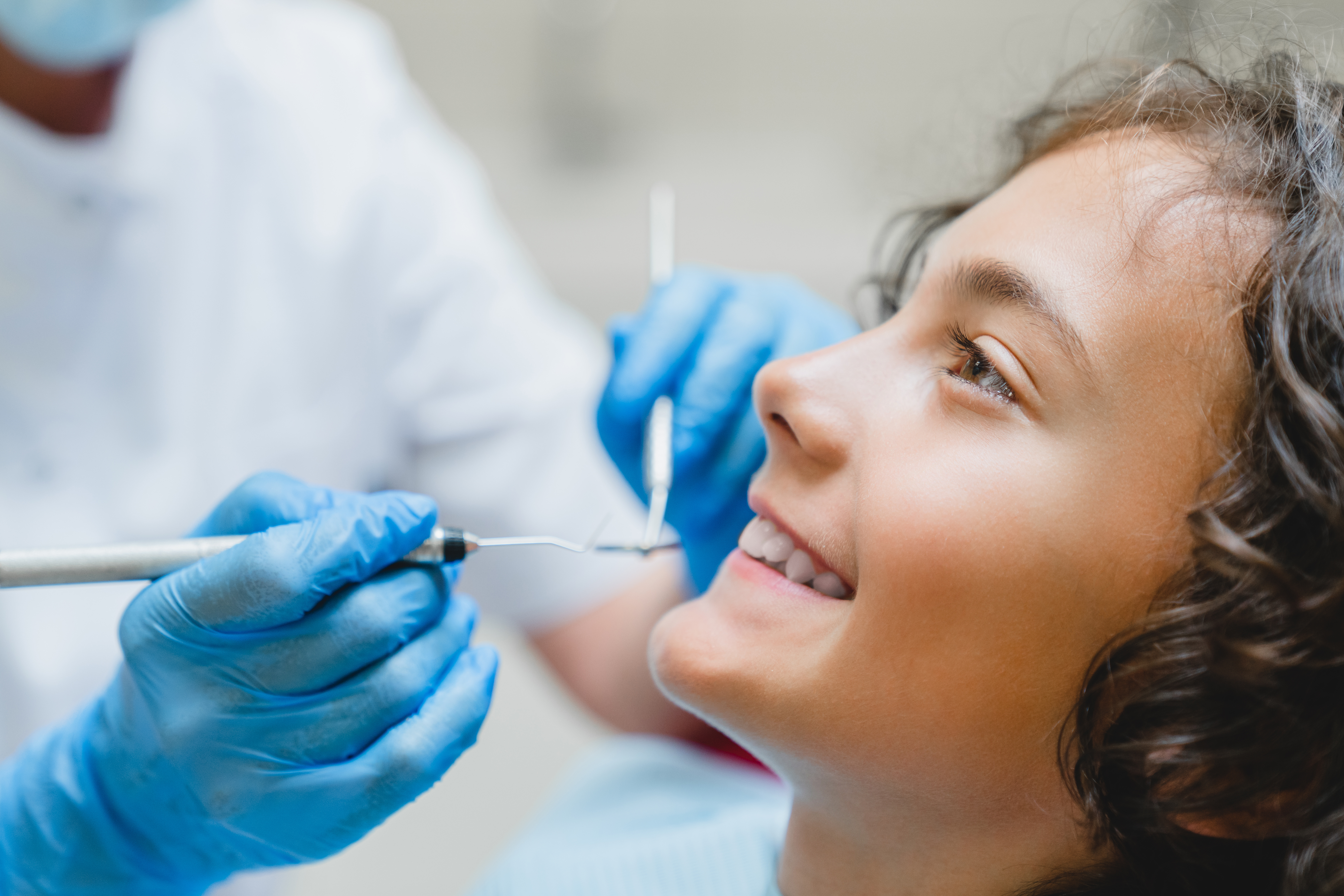 Child smiling at the dentist's office, while the dentist holds tools for cleaning teeth to the child's face