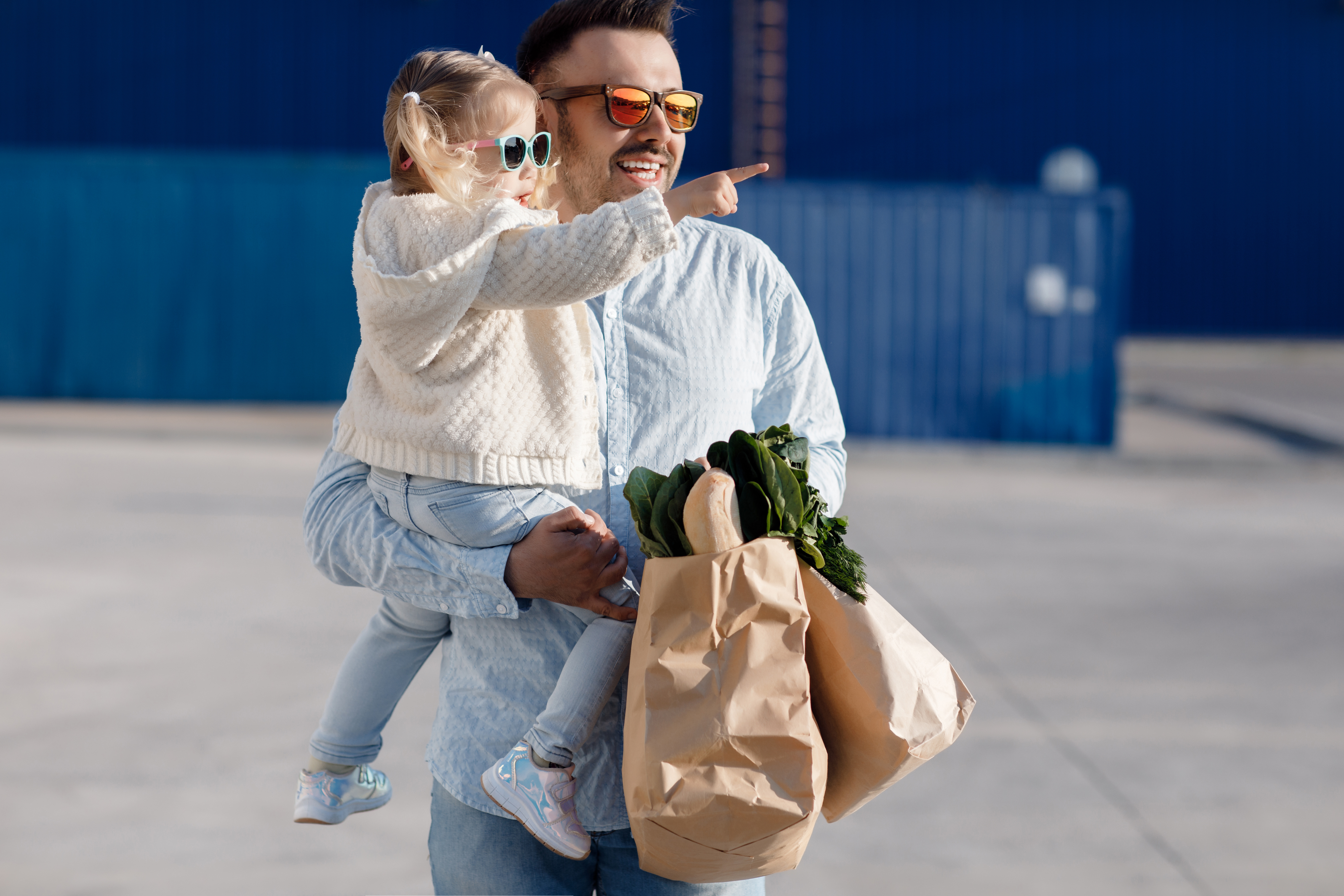 There is a father holding his daughter in a parking lot with sunglasses on while she is pointing at something.