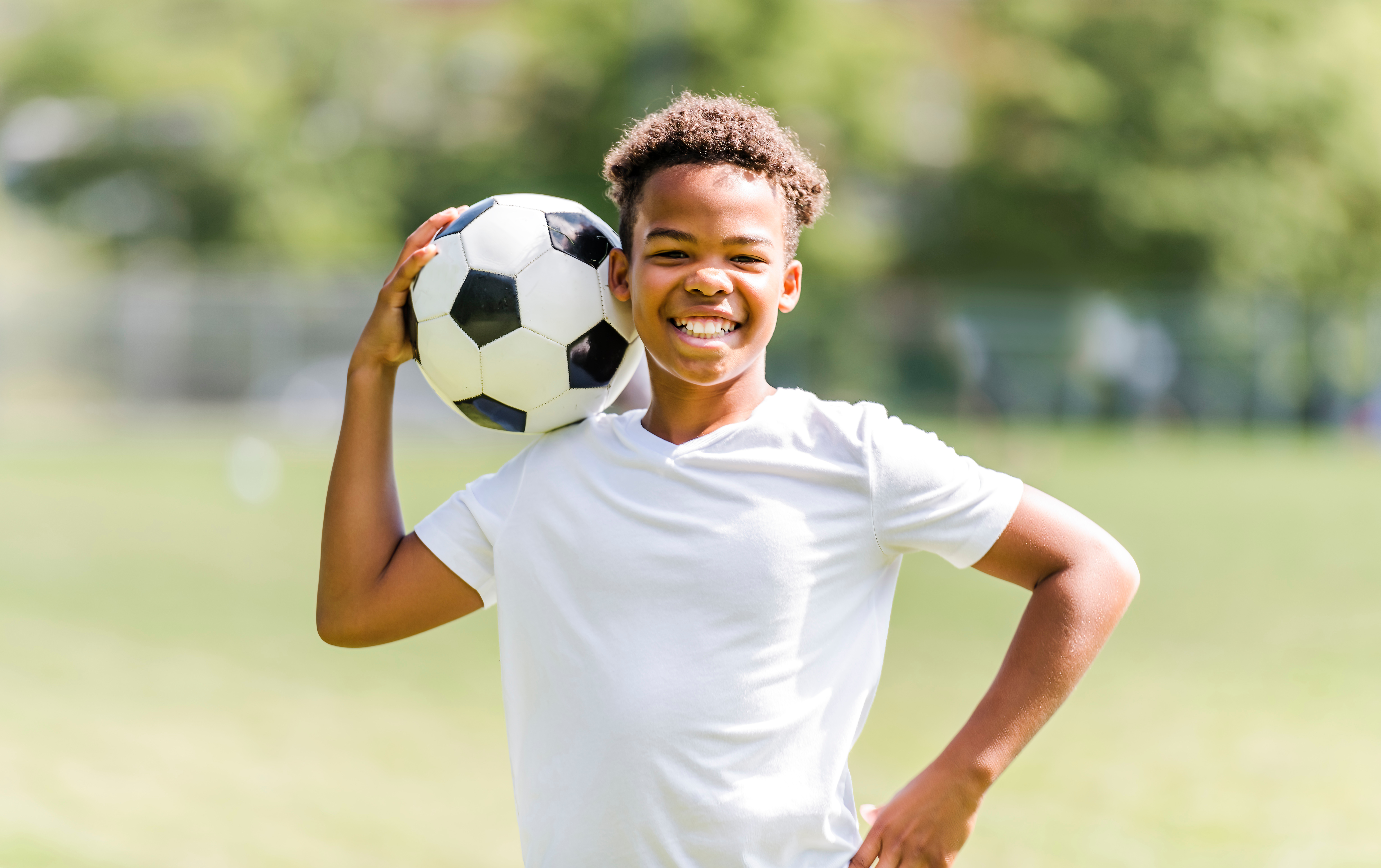 A young boy wearing a white shirt is smiling at the camera while holding a soccer ball.
