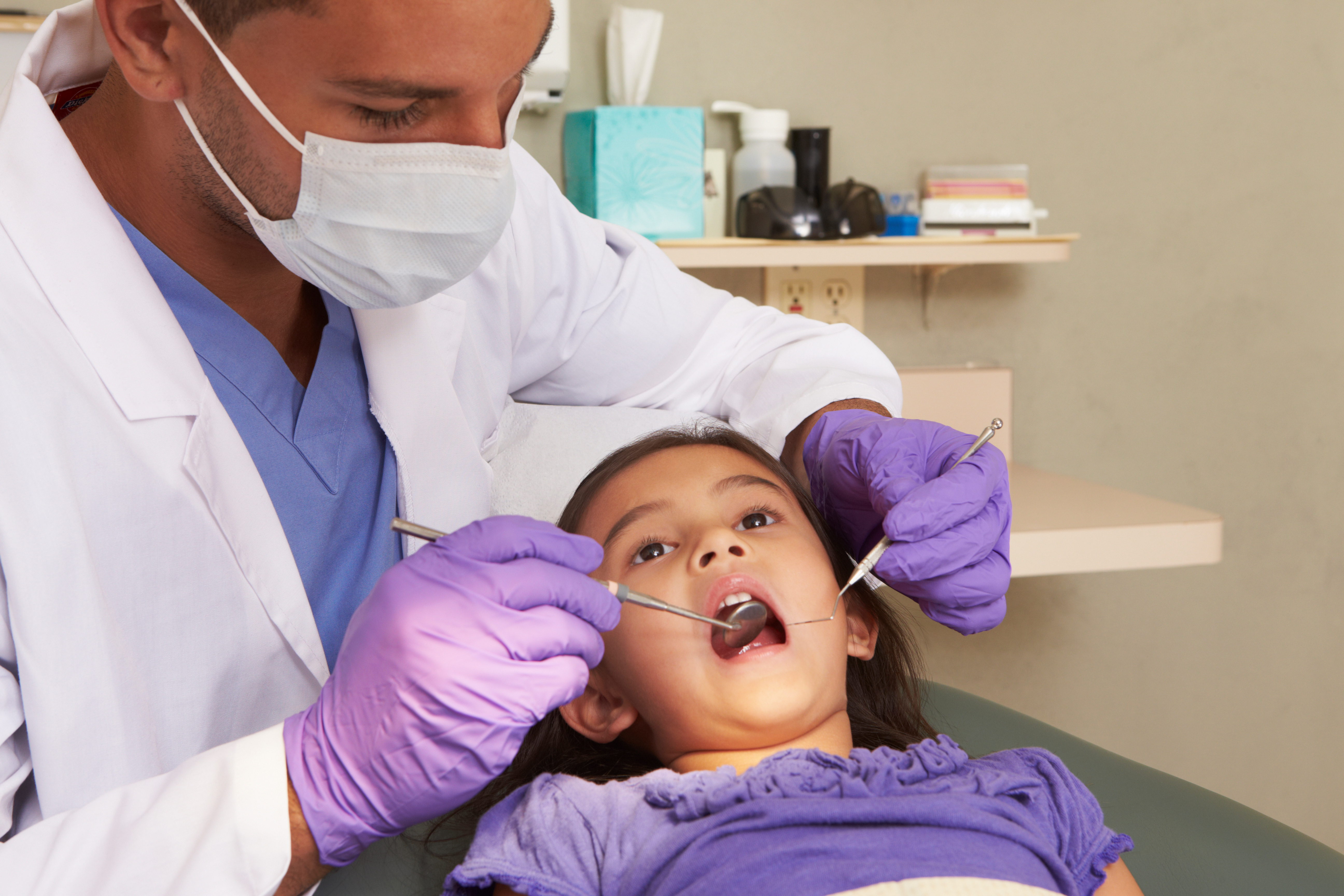 A young patient has their mouth open in a dentist's chair, while the dentist looks into their mouth with tools for an exam.