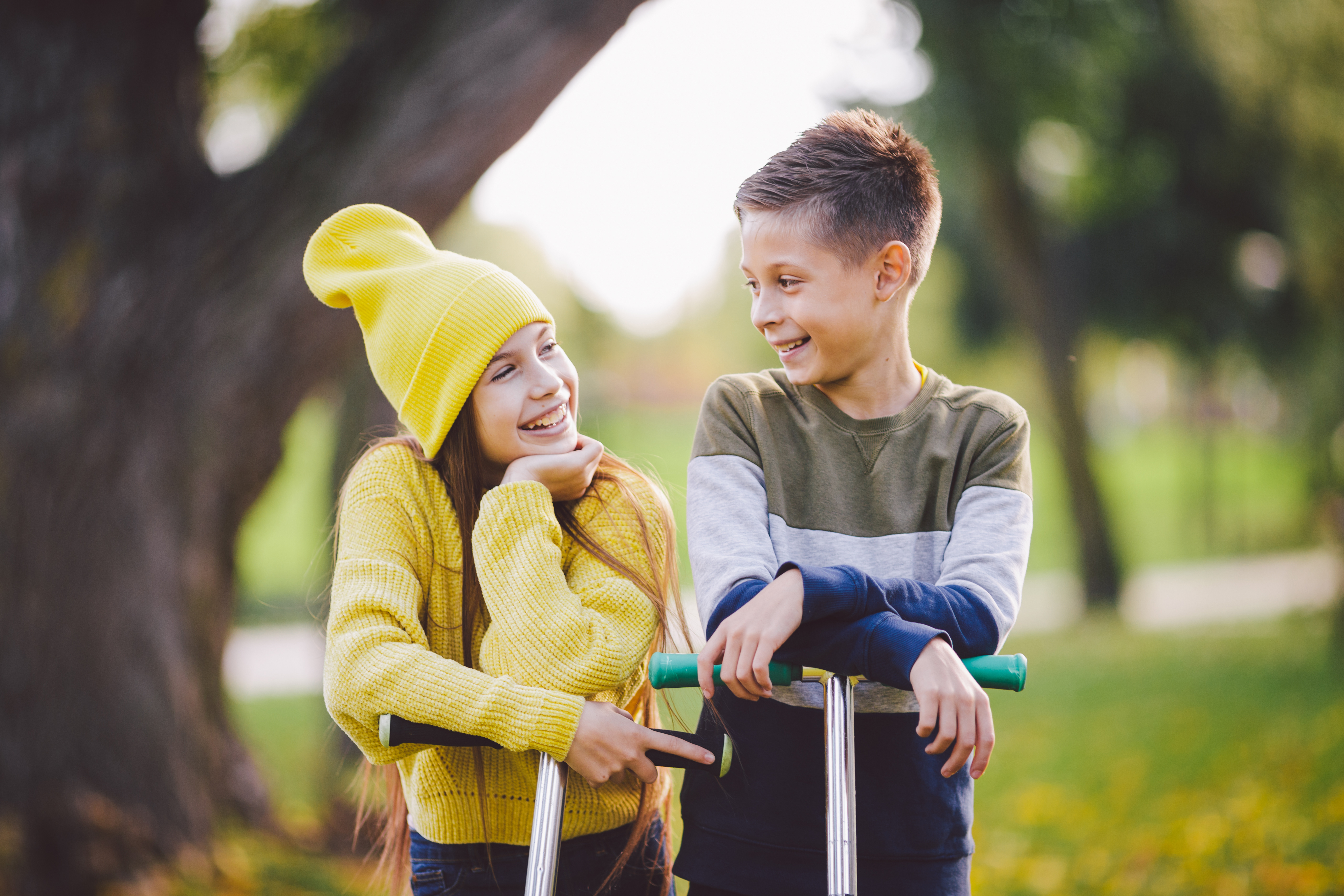 A young girl and young boy are riding their scooters in the fall. The young girl is wearing a knit hat and smiling back at the young boy.