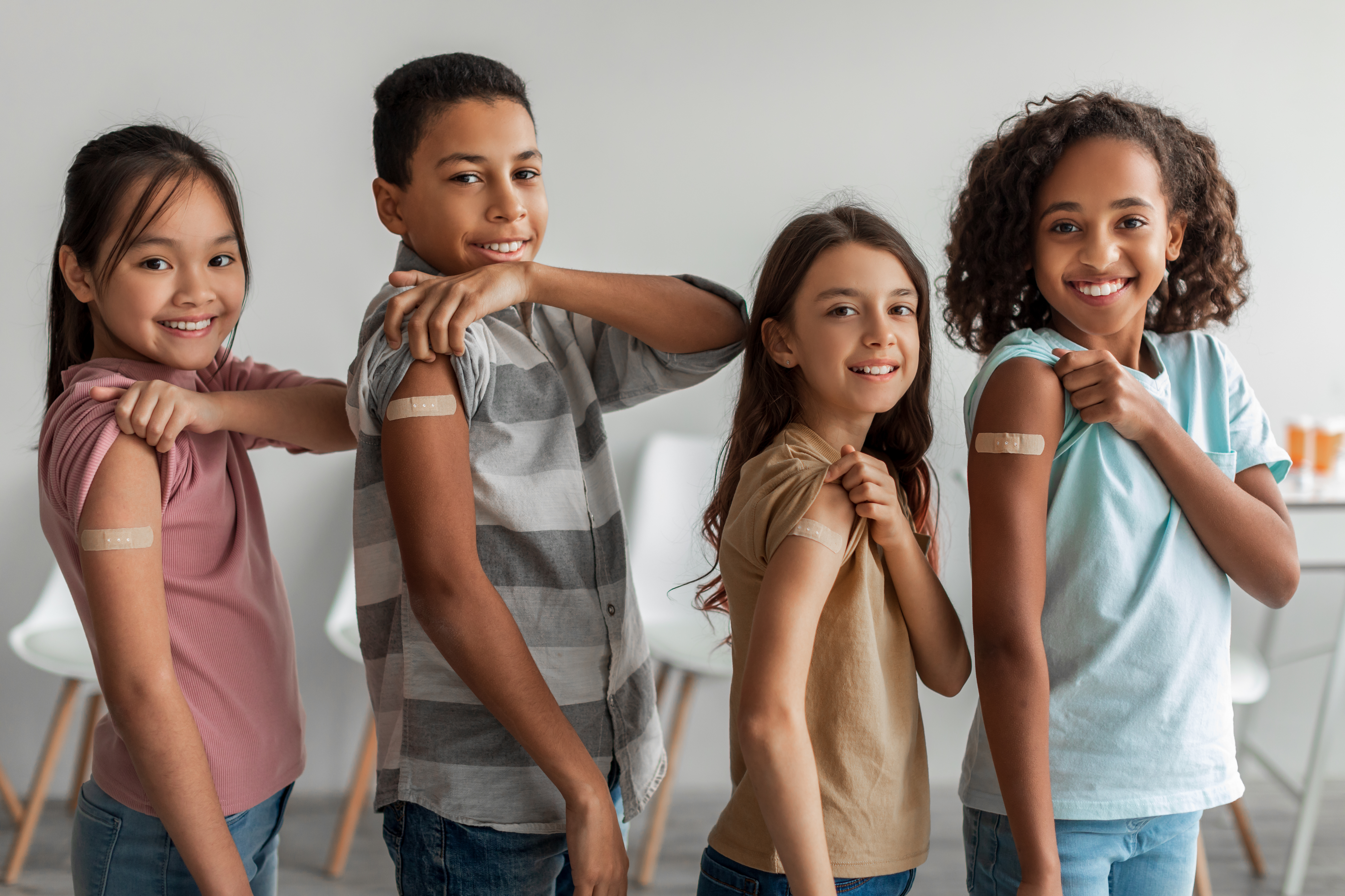 Four smiling children stand in a row, proudly showing their upper arms with bandages after receiving vaccinations. They wear casual clothing and appear confident and happy, promoting a positive message about immunization.