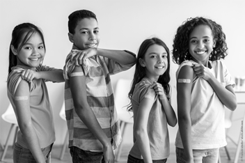 Four smiling children stand in a row, proudly showing their upper arms with bandages after receiving vaccinations. They wear casual clothing and appear confident and happy, promoting a positive message about immunization.