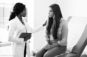 A doctor comforts a teenage girl during a serious medical conversation in an exam room, offering support with a gentle touch.