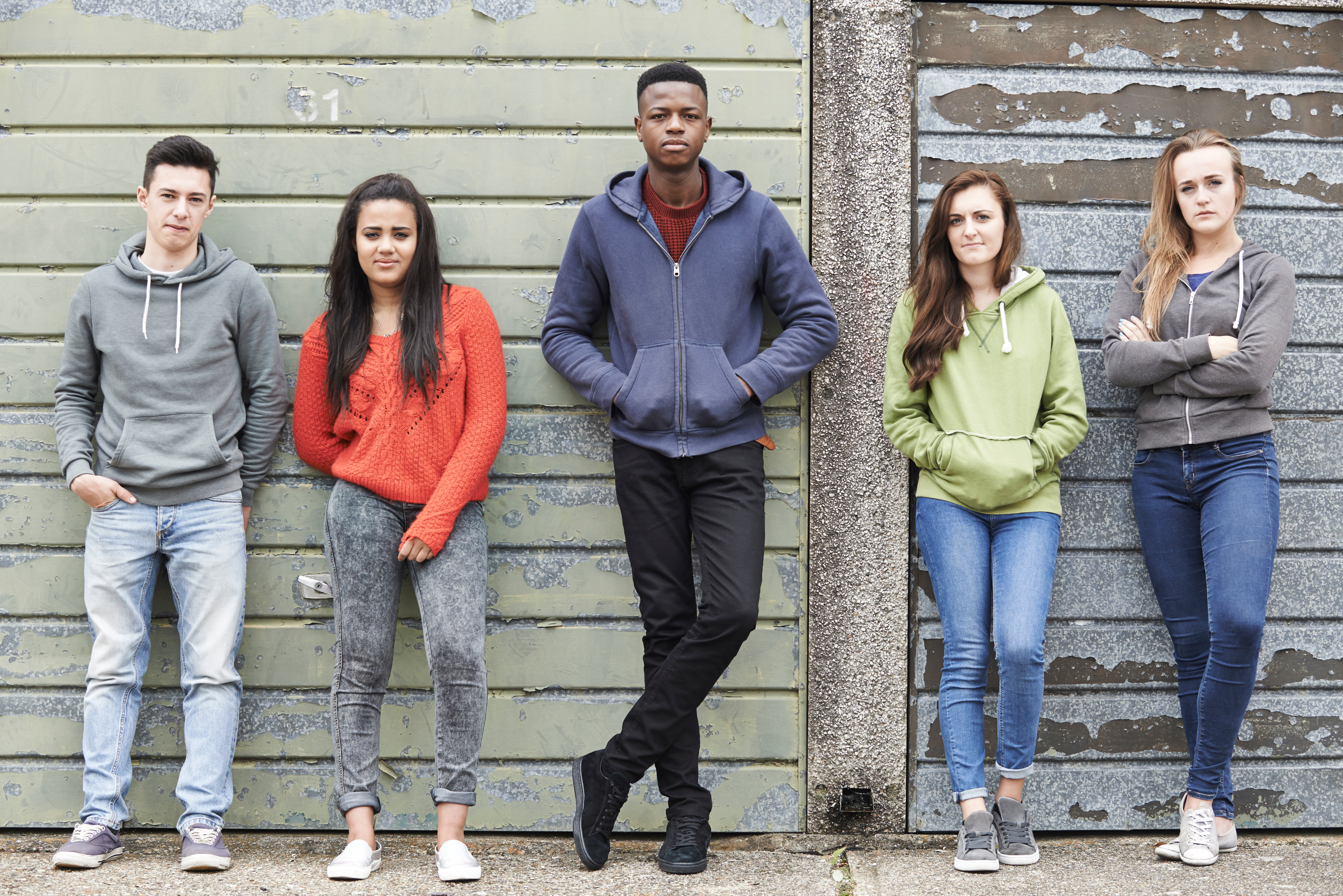 Five teens are leaning against a wall in a line with their hands in their pockets.