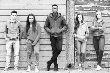 Five teens are leaning against a wall in a line with their hands in their pockets.