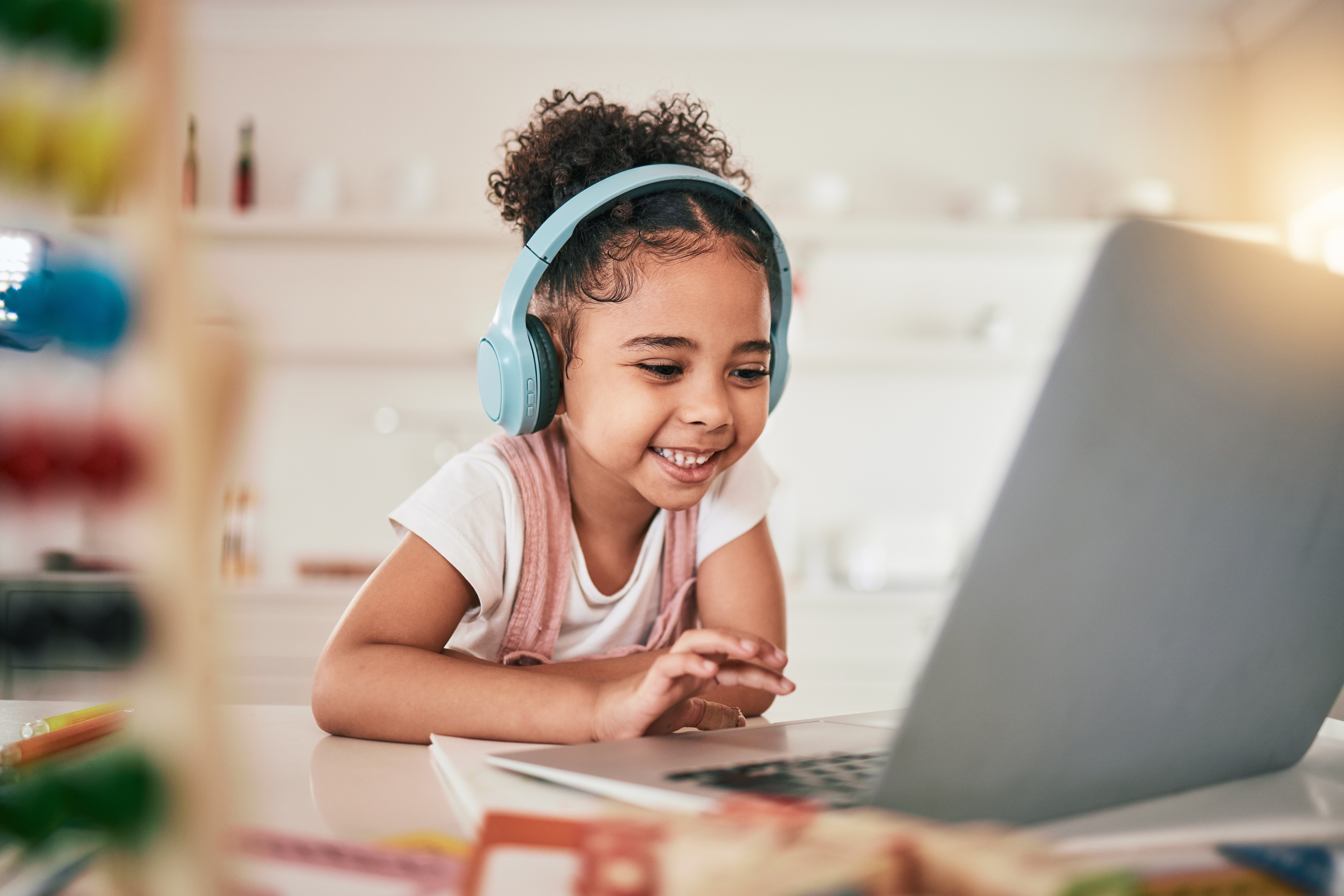 A young girl wearing over-ear headphones smiles while using a laptop at a table. She appears engaged and happy in a bright indoor setting, suggesting a learning or communication activity.