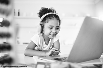 A young girl wearing over-ear headphones smiles while using a laptop at a table. She appears engaged and happy in a bright indoor setting, suggesting a learning or communication activity.
