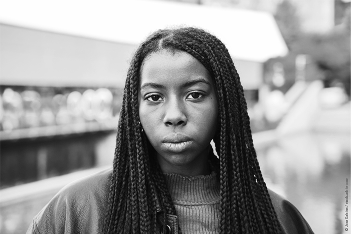A young woman with long braids looks directly into the camera with a serious expression. She is standing outdoors near a body of water, wearing a leather jacket and a ribbed sweater.