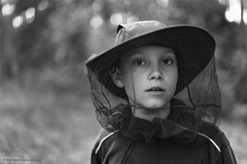 A young child wearing a wide-brimmed hat with mosquito netting looks ahead with a serious expression. He stands in a wooded outdoor area, with soft light filtering through the trees in the background.