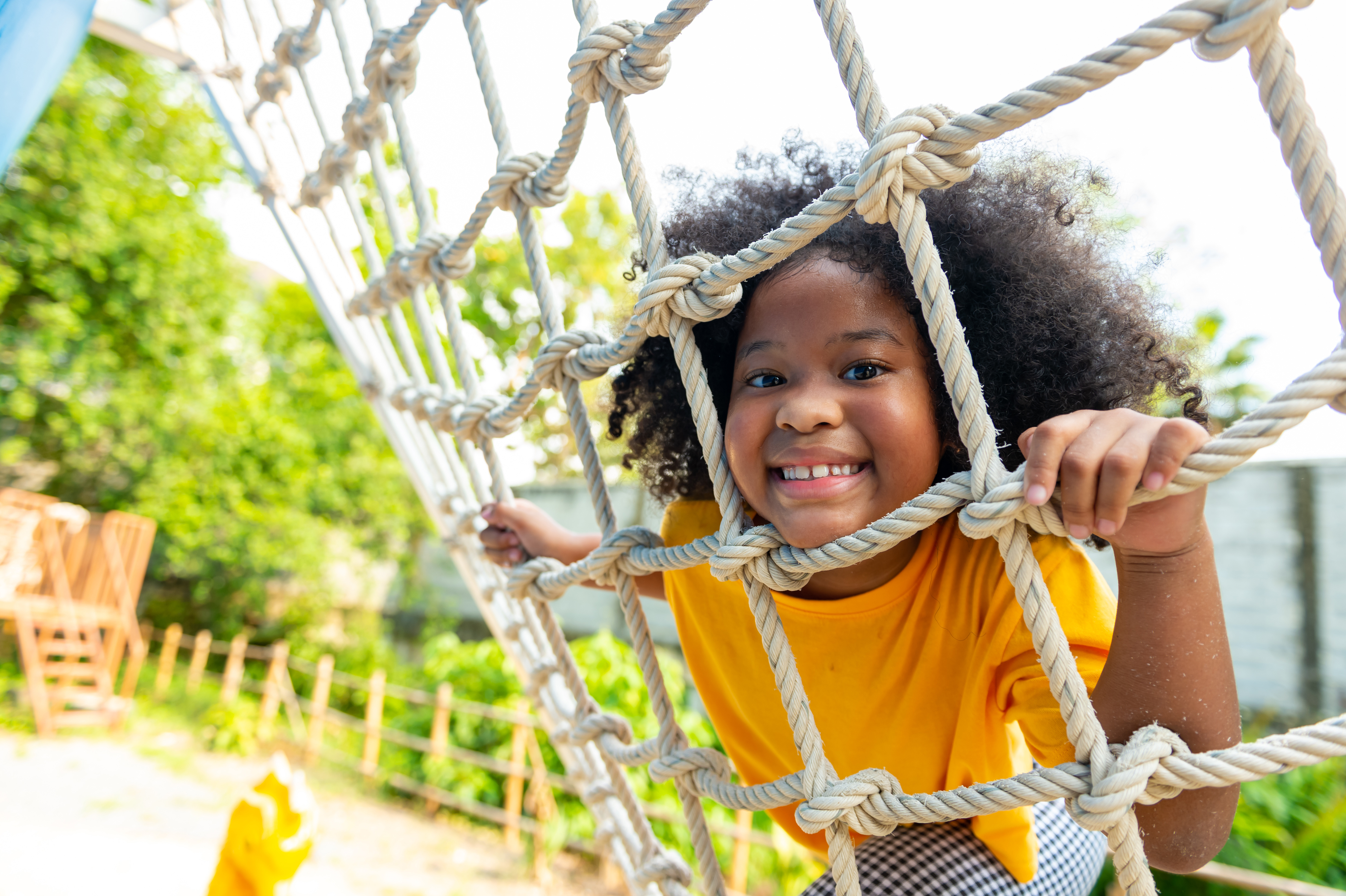 A young girl with curly hair smiles while climbing through a rope net on a playground. She is framed by knotted ropes and looks directly at the camera, appearing happy and active.
