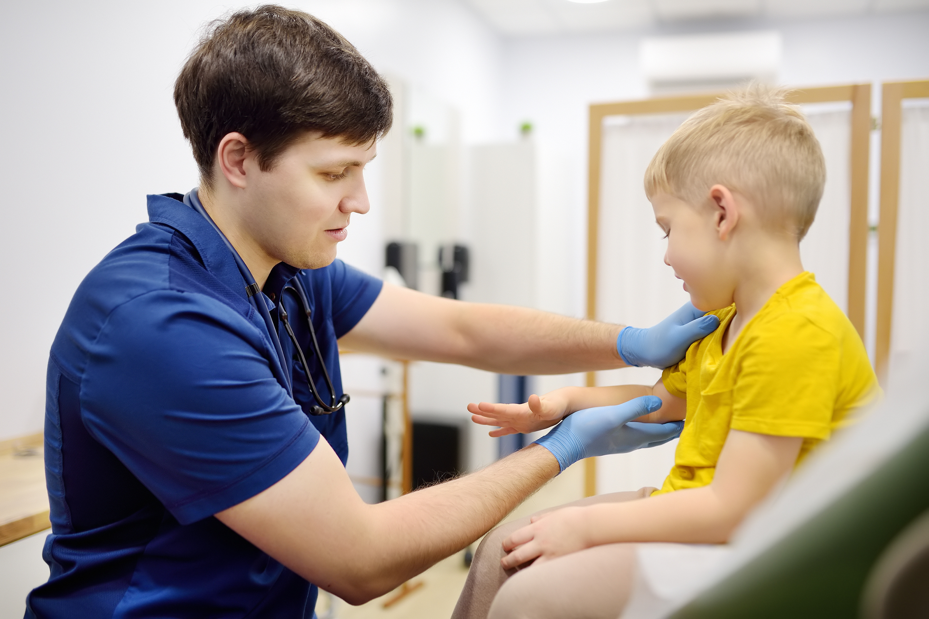 A healthcare professional in a clinical setting examines a child seated on an examination table. The professional is using both hands to check the child's upper chest and arm area. Medical equipment and partitions are visible in the background.