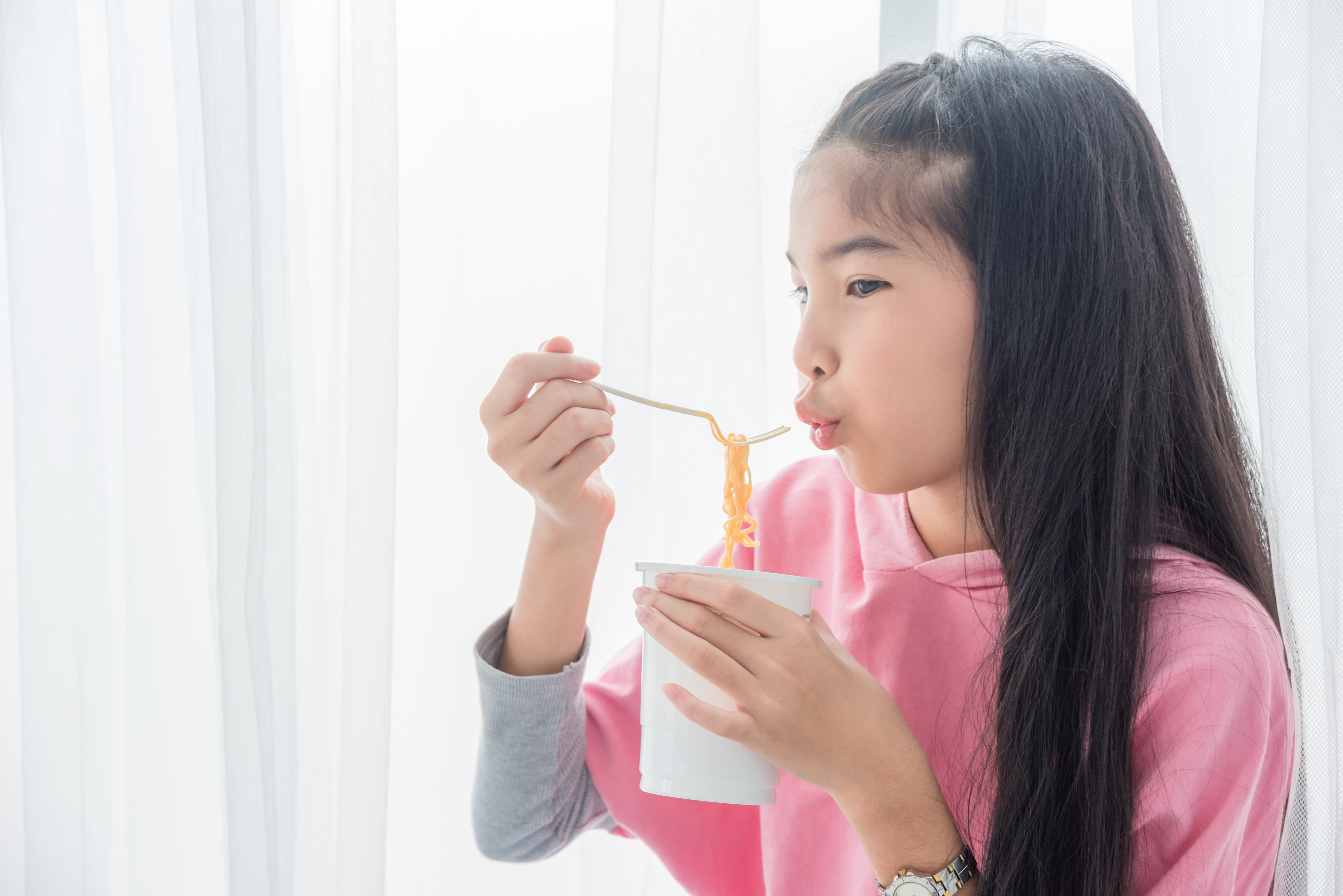A person with long straight hair is holding a white cup in one hand and using a fork to lift noodles from the cup. The image is in black and white, with soft lighting coming from sheer curtains in the background. The person is wearing a long-sleeved top and a wristwatch on the left wrist.