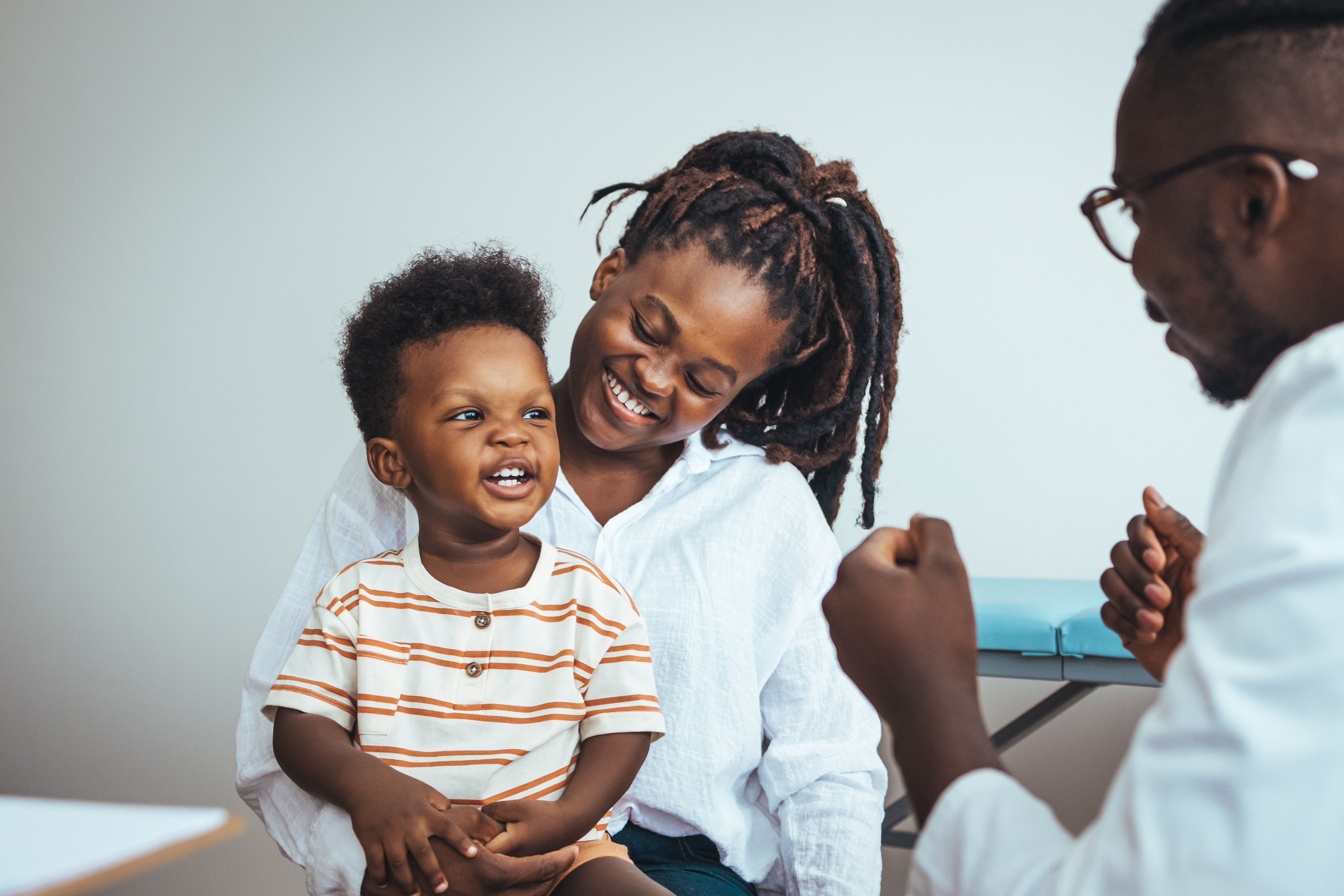 A smiling mother and her young child sit together during a medical appointment. The child sits on the mother’s lap, and both are engaged in a cheerful conversation with a healthcare provider, who is partially visible in the foreground.