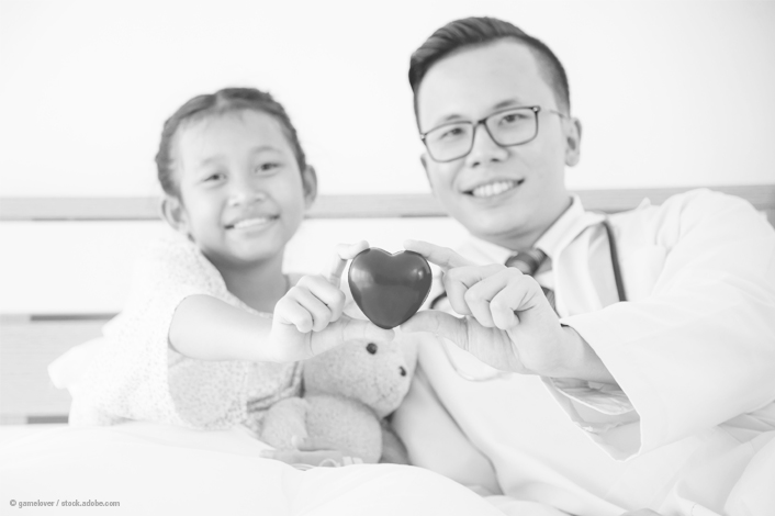 A smiling young patient and a doctor sit side by side, holding a heart-shaped object together. The child, sitting up in a hospital bed and hugging a stuffed animal, and the doctor, wearing a white coat and stethoscope, both look happy and connected