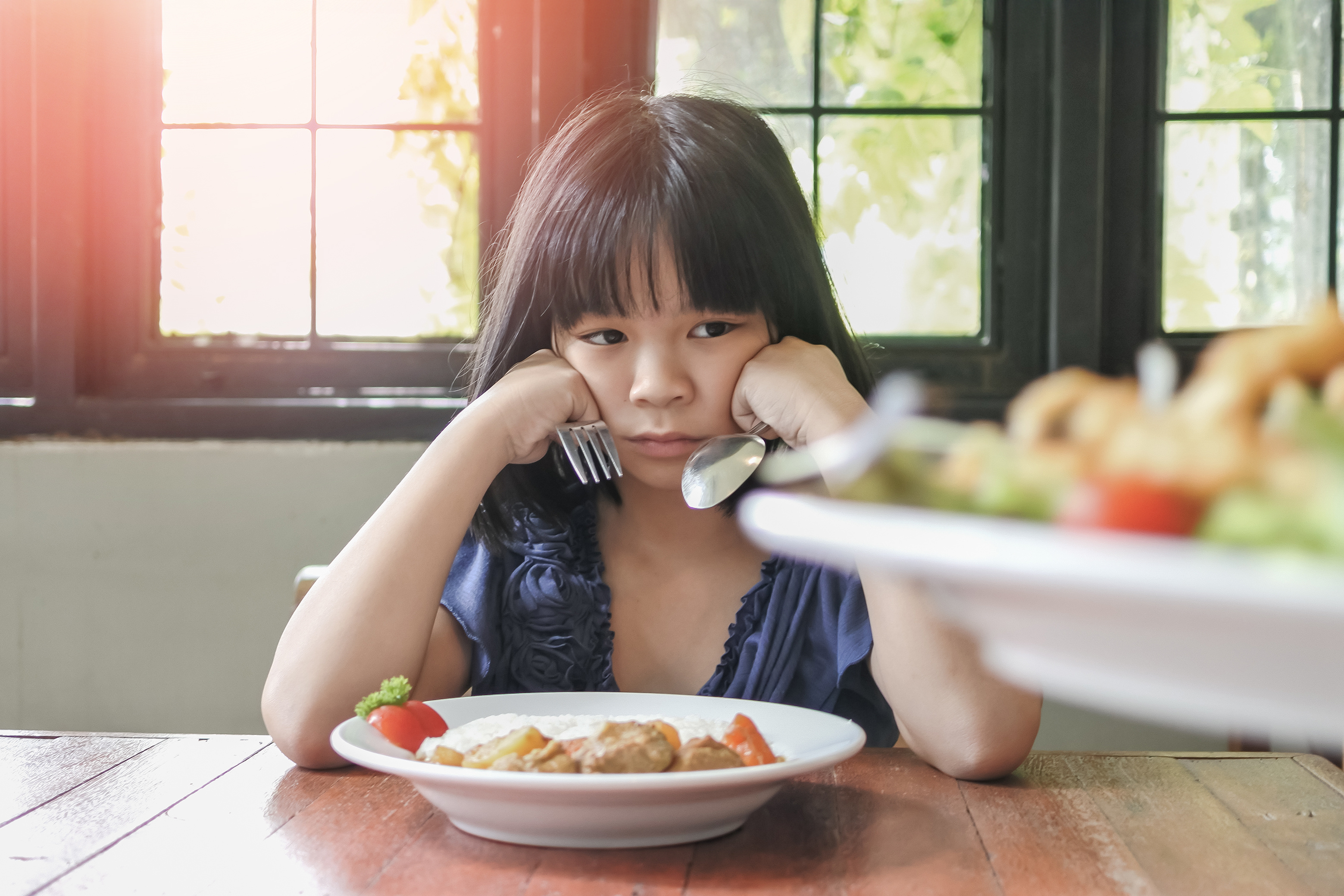 A young girl sits at a table in front of a plate of food, resting her head on her hands while holding a fork and spoon. She looks unhappy and uninterested in the meal, conveying reluctance or discomfort with eating.