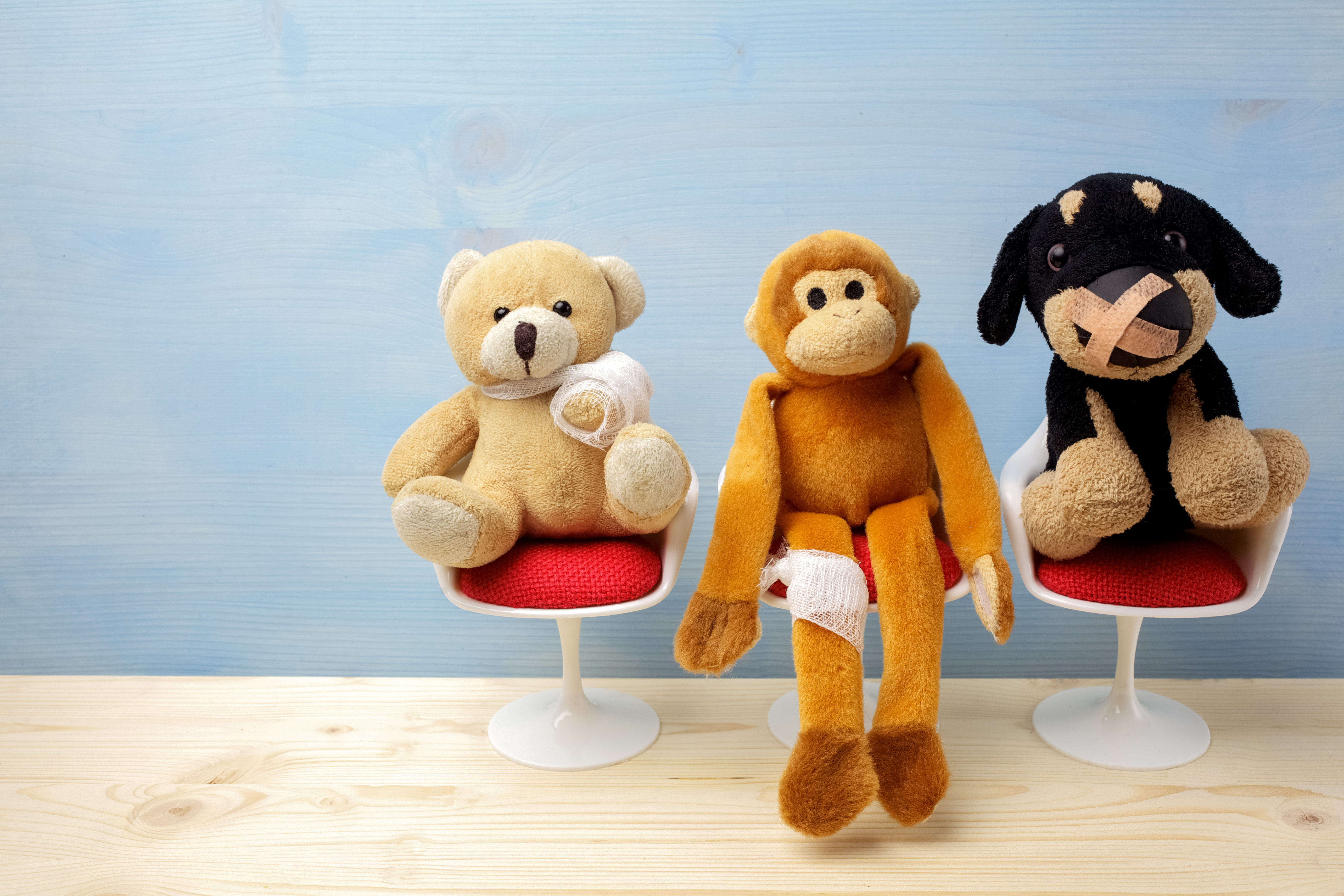 Three stuffed animals—a teddy bear with a bandaged arm, a monkey with a leg wrap, and a dog with a bandaged snout—sit on small white chairs, resembling patients in a waiting room. The scene conveys a playful, child-friendly healthcare environment.